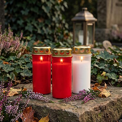 Grabkerzen mit Deckel: zwei rote und ein weißes Grablicht mit goldfarbenem Lochdeckel auf einer Grabplatte zwischen Heidekraut und Efeu